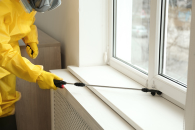 Photo of Pest control worker in protective suit spraying pesticide near window indoors, closeup