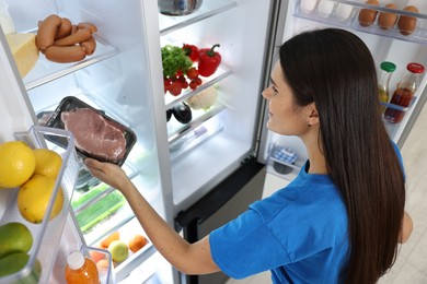 Photo of Young woman taking pack of meat out of refrigerator indoors, above view