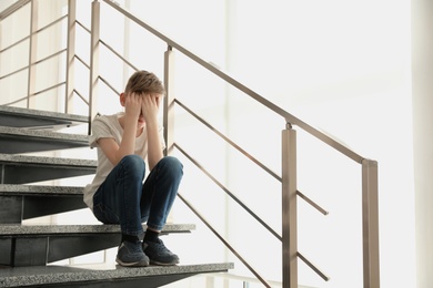 Sad little boy sitting on stairs indoors. Space for text