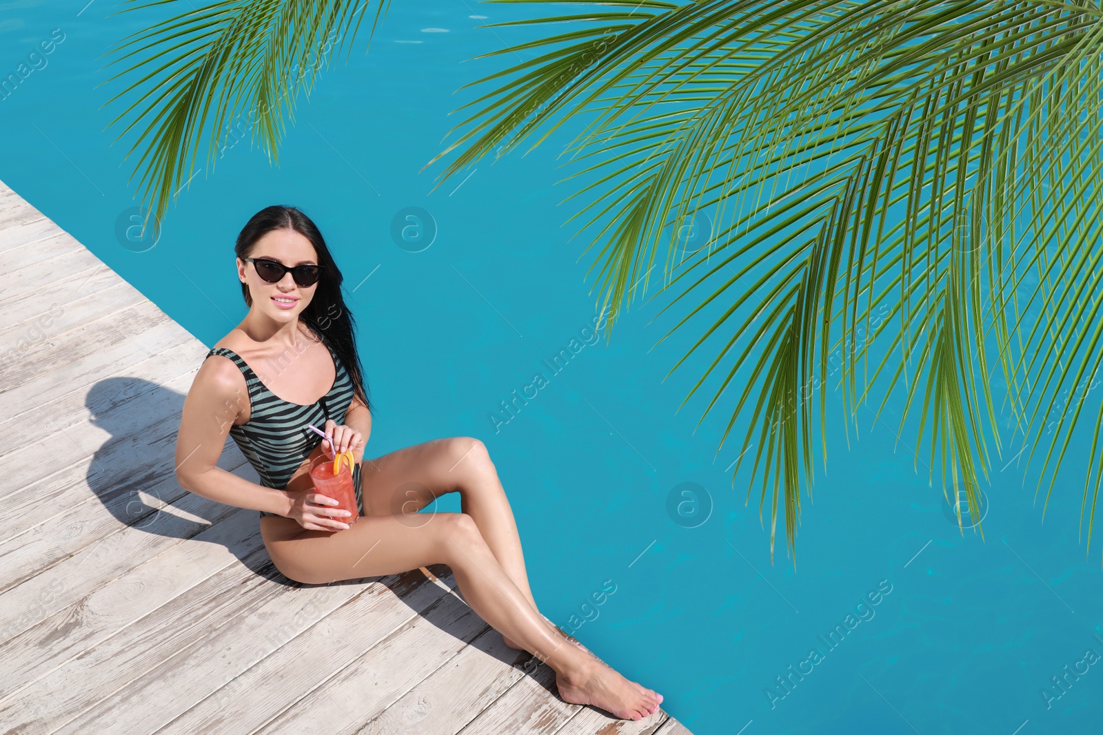 Image of View of beautiful green tropical leaves and young woman with refreshing cocktail near swimming pool on sunny day, above view