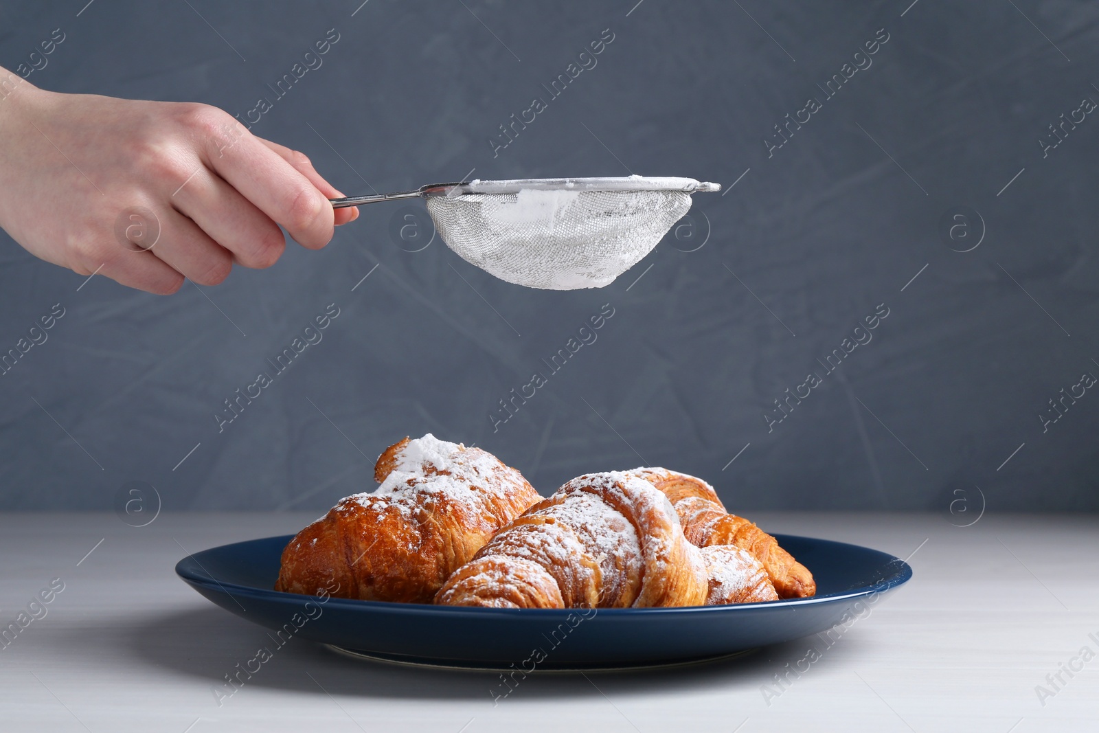 Photo of Woman with sieve sprinkling powdered sugar onto croissants at white wooden table, closeup