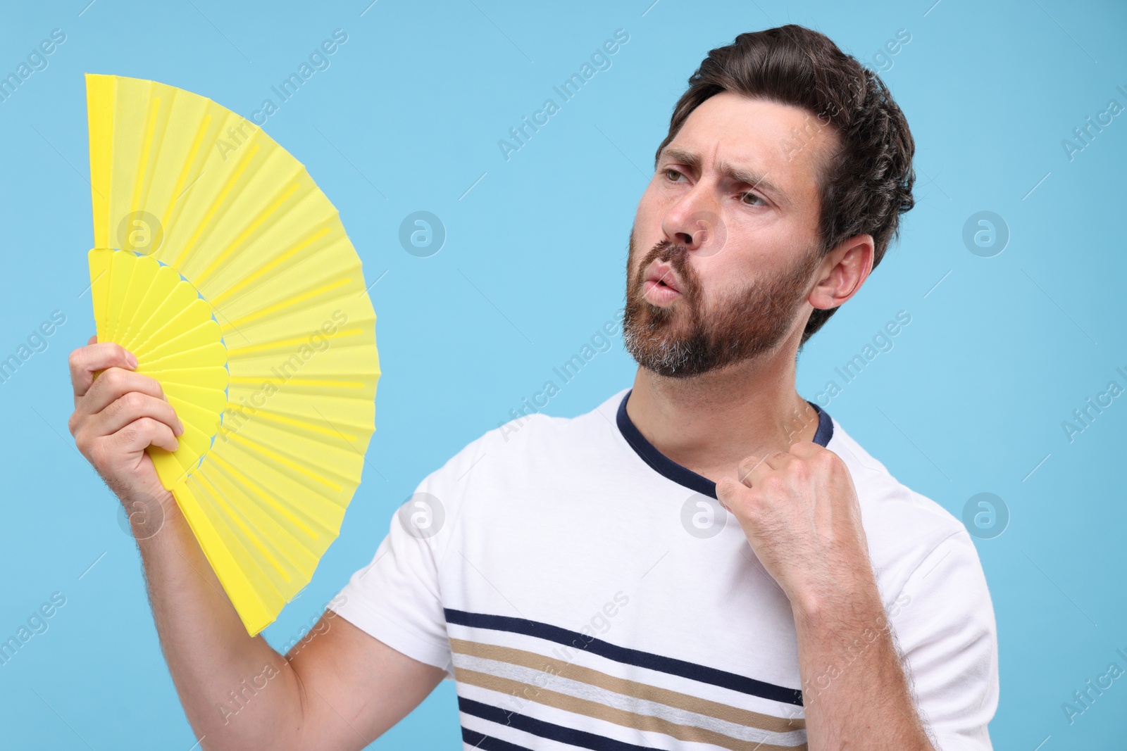 Photo of Unhappy man with hand fan suffering from heat on light blue background