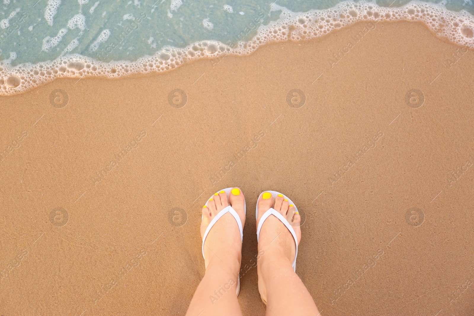 Photo of Top view of woman with white flip flops on sand near sea, space for text. Beach accessories