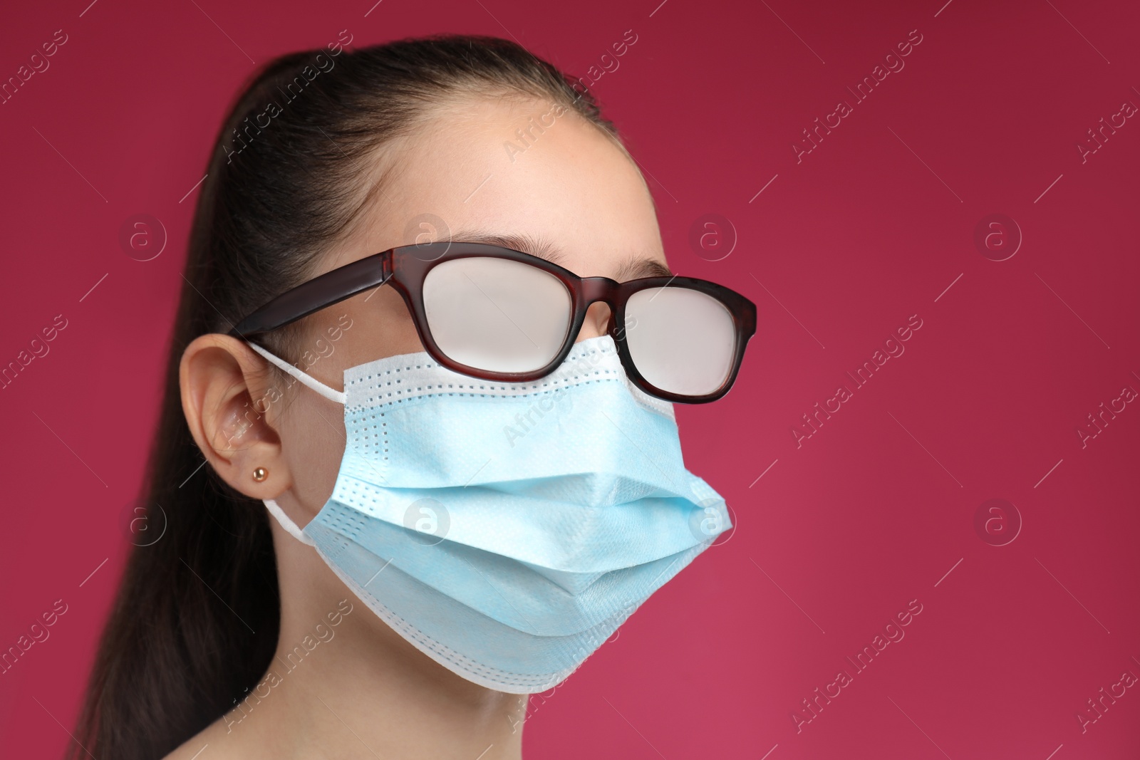 Photo of Little girl with foggy glasses caused by wearing medical face mask on pink background. Protective measure during coronavirus pandemic