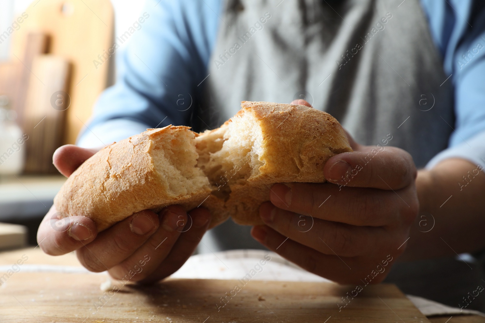 Photo of Man breaking loaf of fresh bread at wooden table indoors, closeup