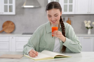 Photo of Happy young woman with glass of juice writing in notebook at white marble table indoors