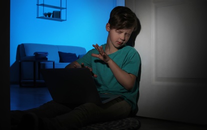 Photo of Frightened little child with laptop on floor in dark room. Danger of internet