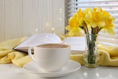 Beautiful yellow daffodils in vase, cup of coffee, book and festive lights on windowsill. Space for text