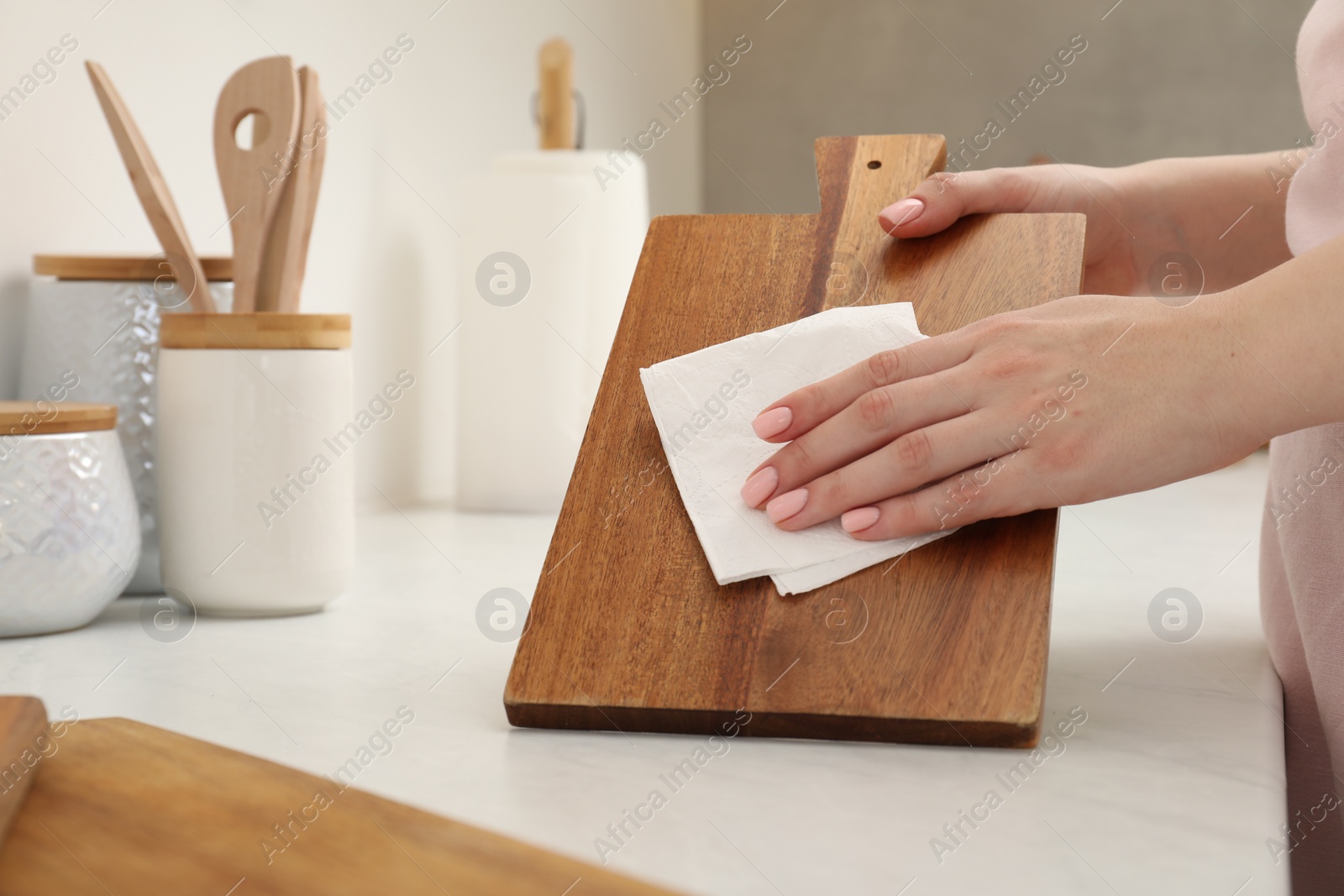 Photo of Woman wiping wooden cutting board with paper napkin at white table, closeup