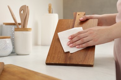 Woman wiping wooden cutting board with paper napkin at white table, closeup