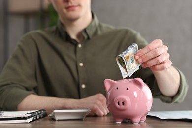 Photo of Financial savings. Man putting dollar banknote into piggy bank at wooden table, closeup