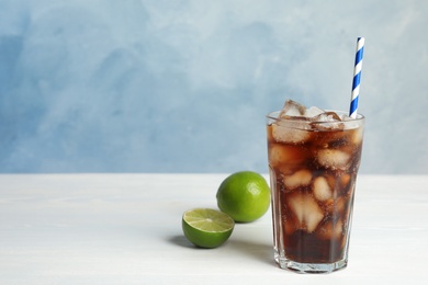Photo of Glass of refreshing soda drink with ice cubes, limes and straw on white wooden table against blue background. Space for text