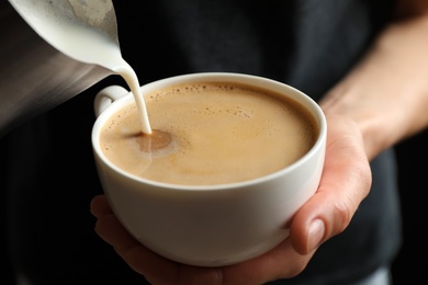 Photo of Woman pouring milk into cup of hot coffee, closeup