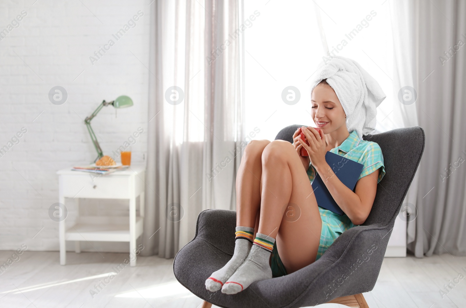 Photo of Young happy woman with book and cup of aromatic coffee in armchair at home. Lazy morning