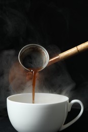 Photo of Turkish coffee. Pouring brewed beverage from cezve into cup against black background