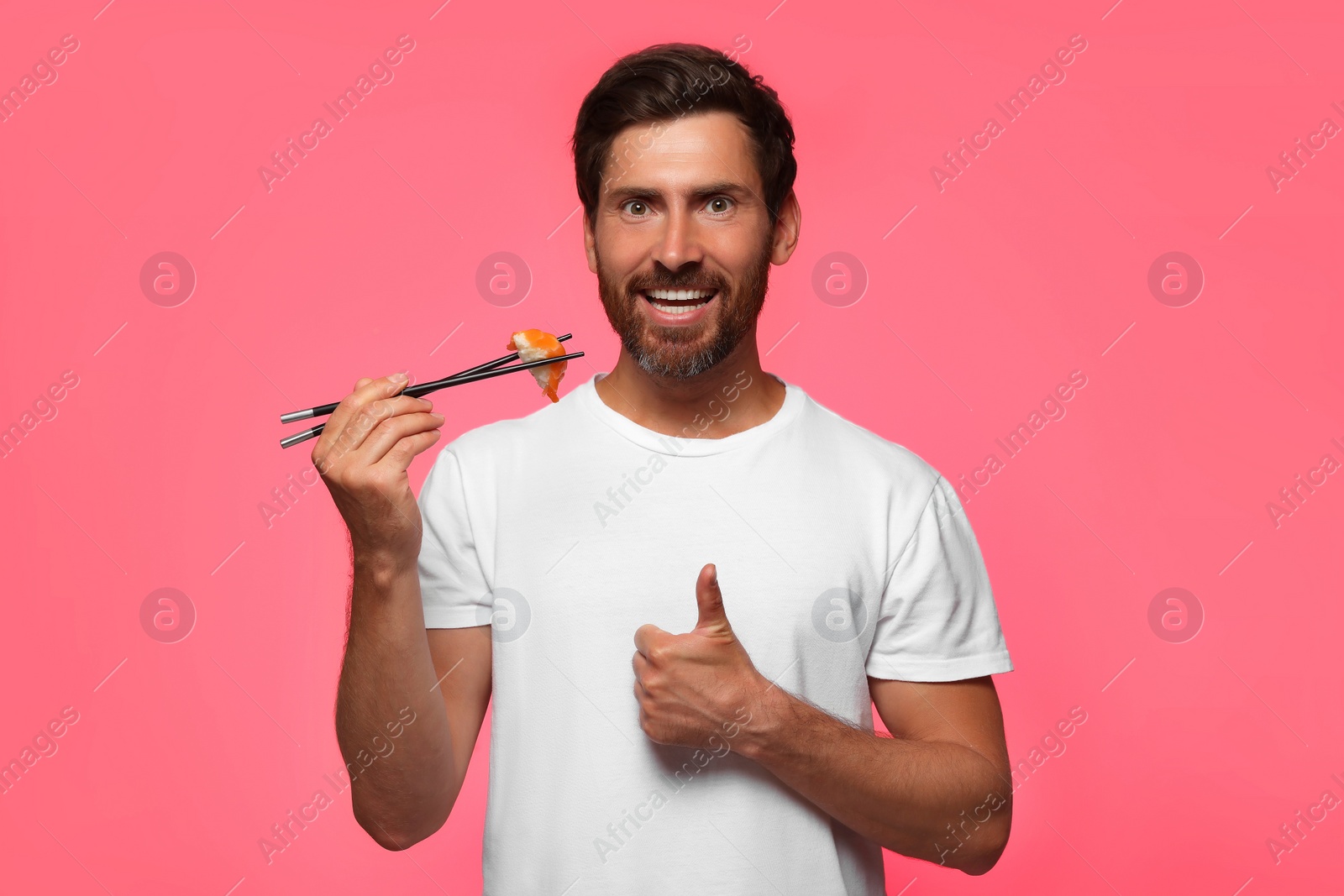 Photo of Happy man holding tasty sushi with chopsticks and showing thumbs up on pink background