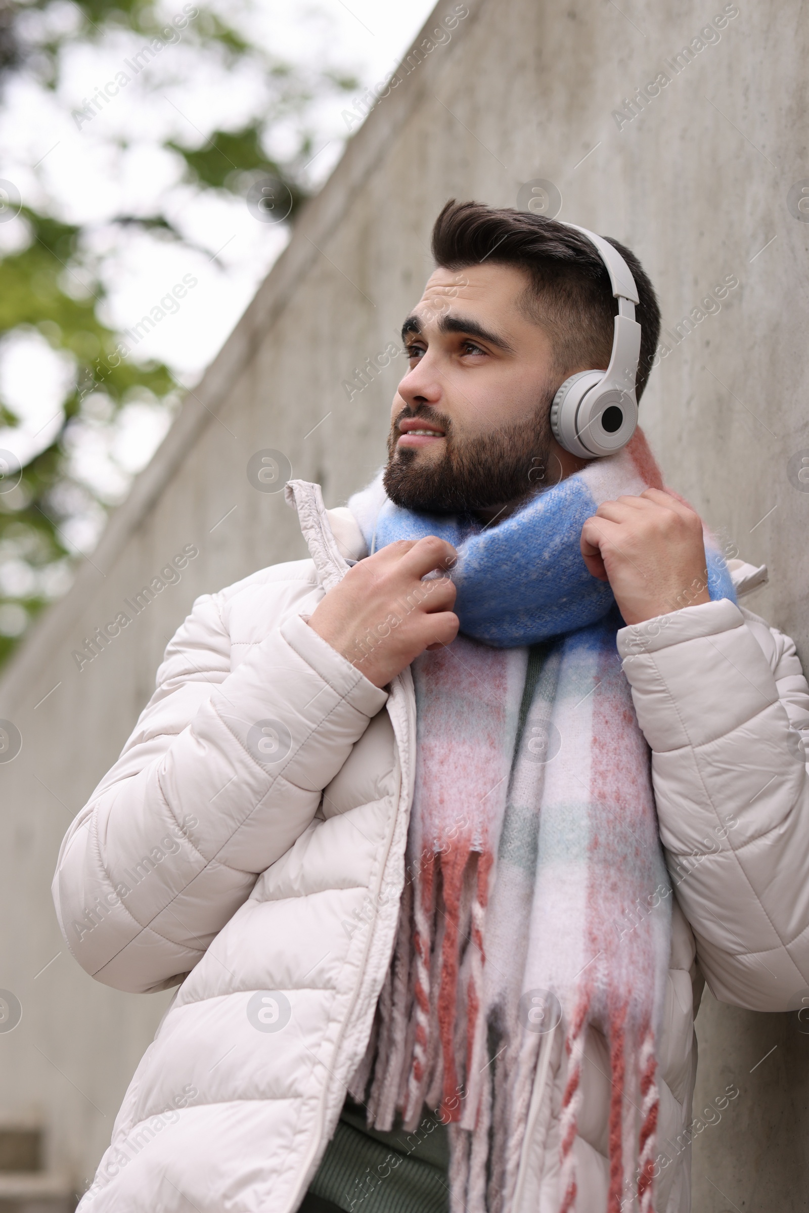 Photo of Handsome man in warm scarf and headphones near wall outdoors