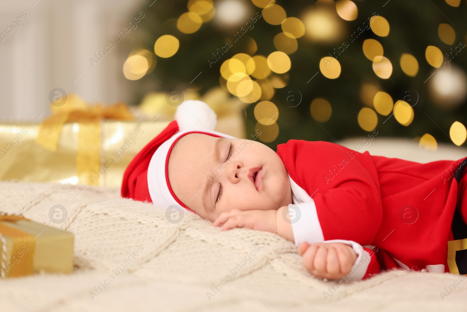 Photo of Cute baby in Christmas costume sleeping on knitted blanket against blurred festive lights. Winter holiday