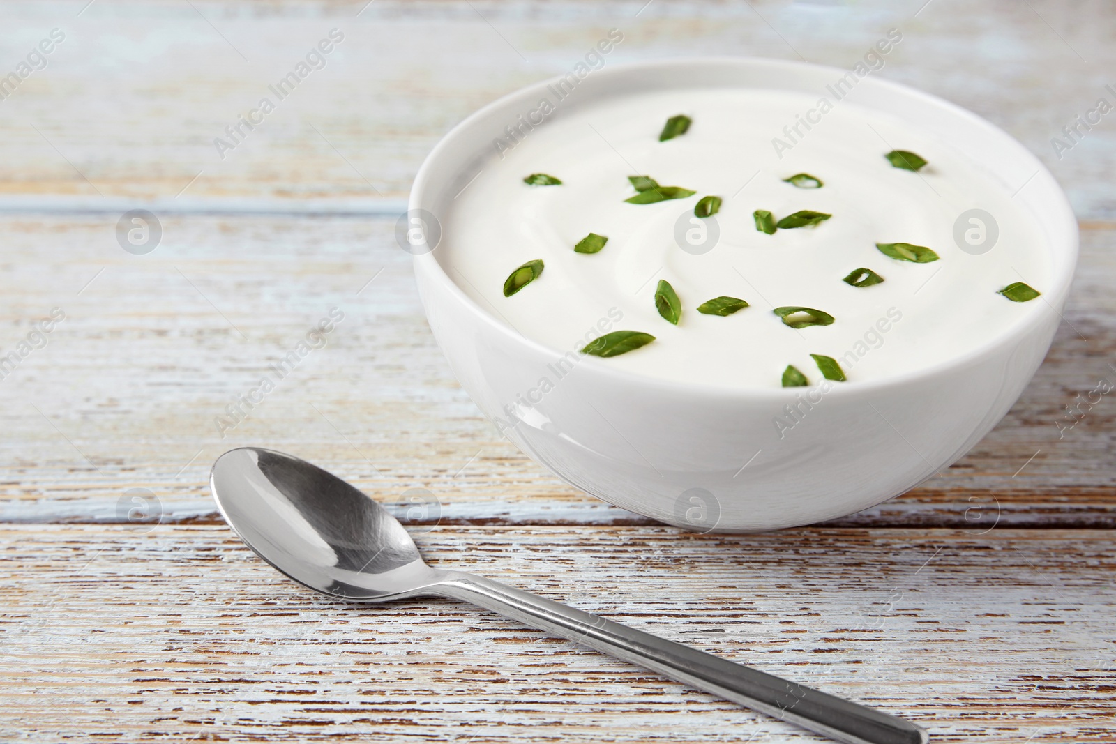 Photo of Bowl of fresh sour cream with green onion and spoon on light blue wooden table. Space for text