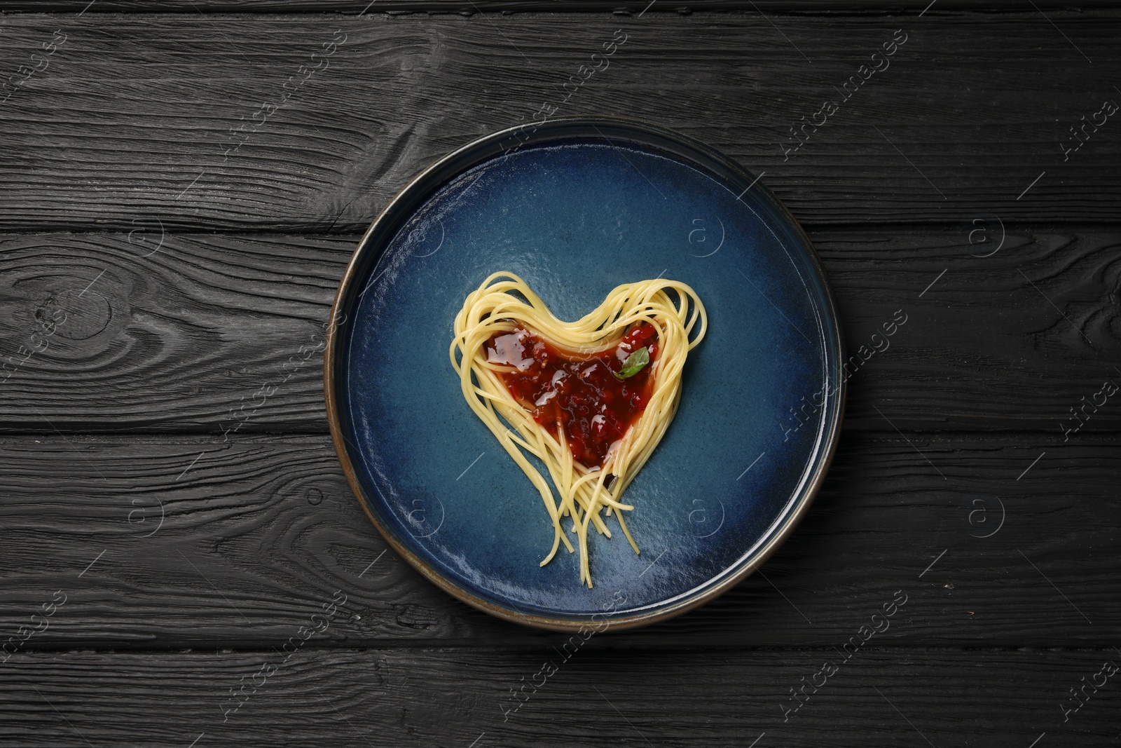 Photo of Heart made with spaghetti and sauce on black wooden table, top view.