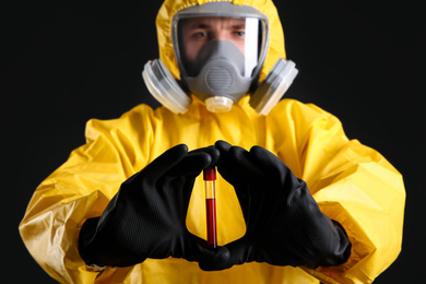 Man in chemical protective suit holding test tube of blood sample against black background, focus on hands. Virus research
