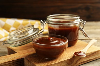 Tasty barbeque sauce in bowl, jar and spoon on wooden table, closeup