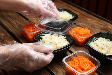 Waiter in gloves closing containers with salads at wooden table, closeup. Food delivery service