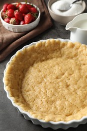 Photo of Making shortcrust pastry. Raw dough in baking dish, strawberries, sugar and milk on grey table