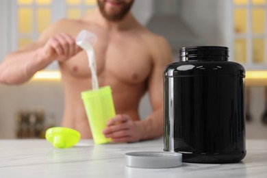Photo of Young muscular man making protein shake at white marble table in kitchen, focus on jar