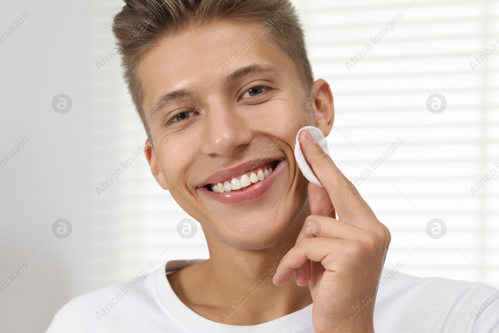 Photo of Handsome young man cleaning face with cotton pad indoors