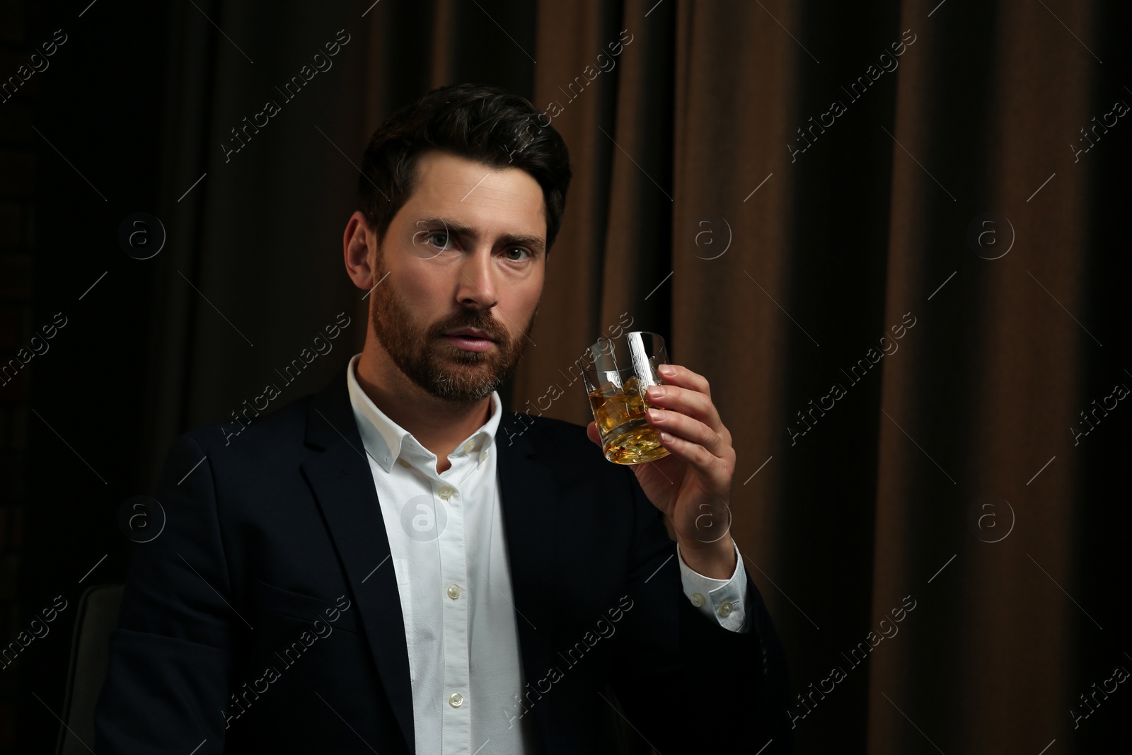 Photo of Man in suit holding glass of whiskey with ice cubes on brown background