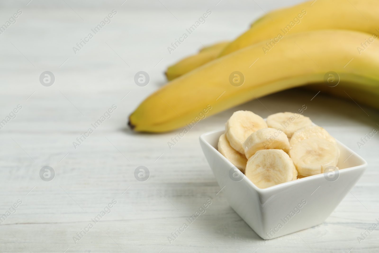 Photo of Bowl with cut bananas near whole fruits on white wooden table. Space for text