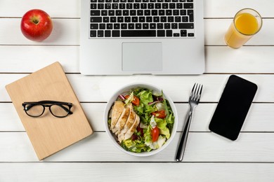 Photo of Bowl with tasty food, glass of juice, laptop, apple, book and smartphone on white wooden table, flat lay. Business lunch