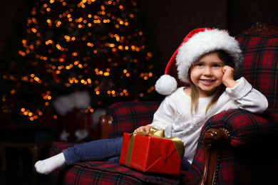 Photo of Cute little child with Christmas gift sitting in armchair at home