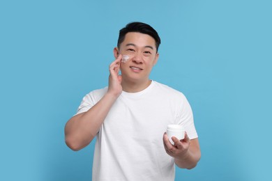 Handsome man applying cream onto his face on light blue background