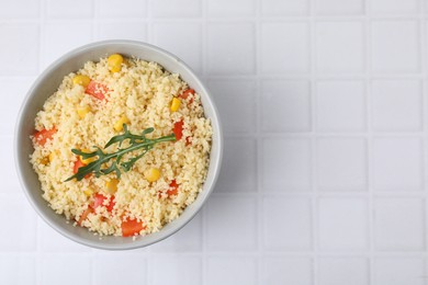 Photo of Tasty couscous with pepper, corn and arugula in bowl on white tiled table, top view. Space for text