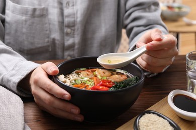 Woman eating delicious ramen with spoon at wooden table, closeup. Noodle soup