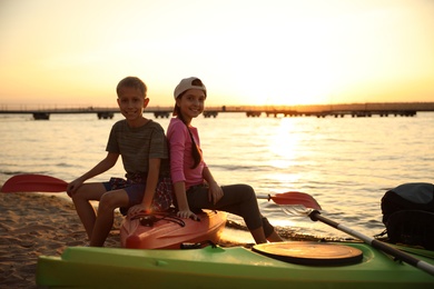 Happy children sitting on kayak near river at sunset. Summer camp