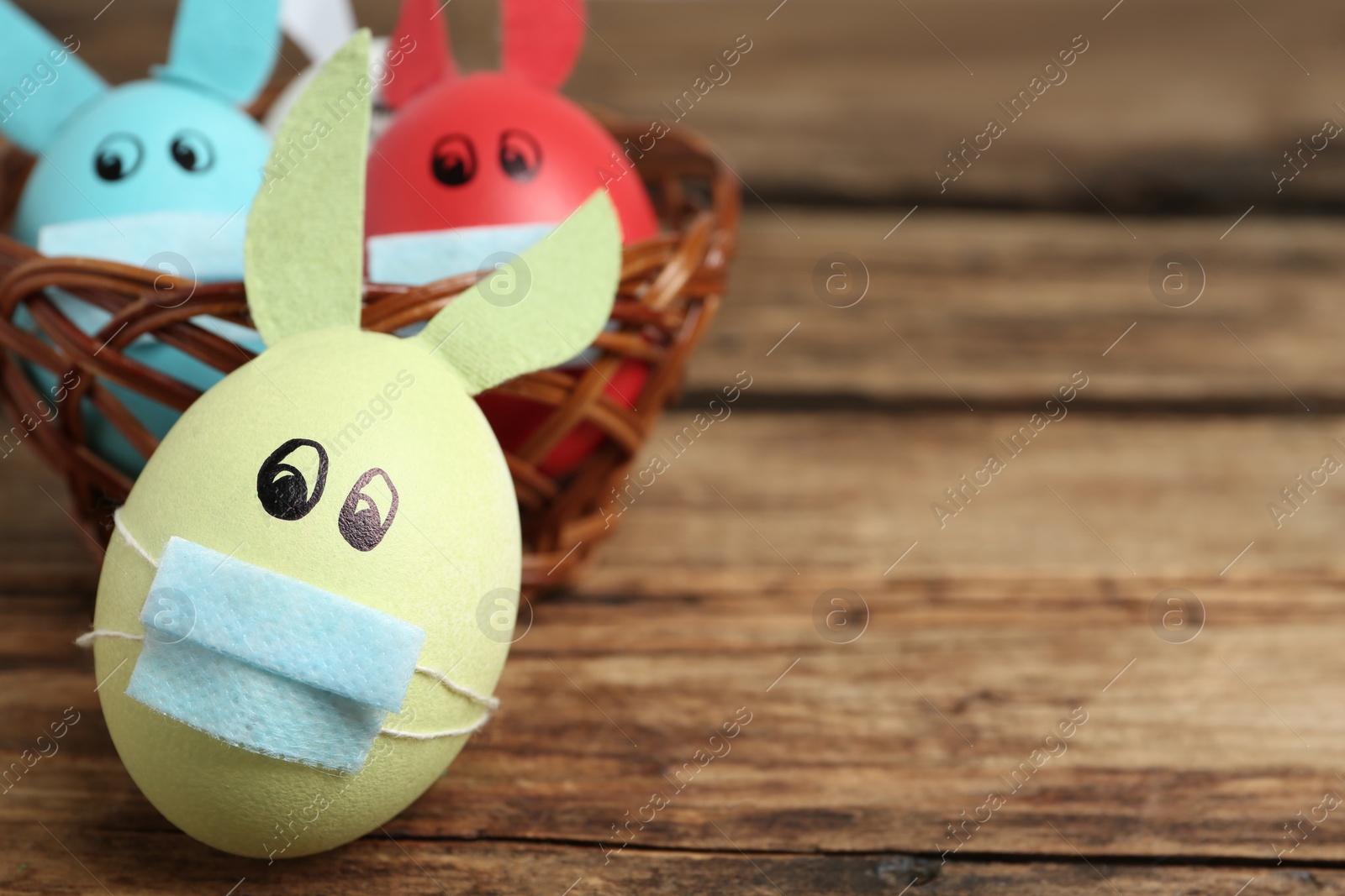 Photo of Painted eggs decorated with bunny ears, protective masks and space for text on wooden table, closeup. Easter holiday during COVID-19 quarantine