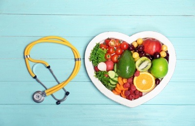 Photo of Heart shaped plate with fresh fruits, vegetables and stethoscope on wooden background, top view. Cardiac diet