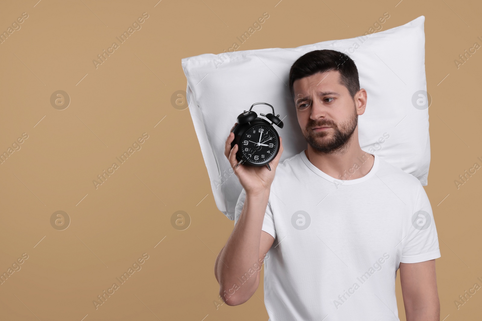 Photo of Sleepy man with pillow and alarm clock on beige background, space for text. Insomnia problem