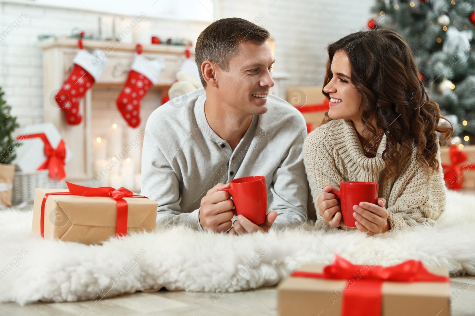 Image of Happy couple with cups of hot drink on floor in room decorated for Christmas