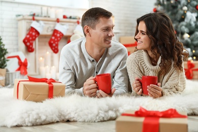 Image of Happy couple with cups of hot drink on floor in room decorated for Christmas