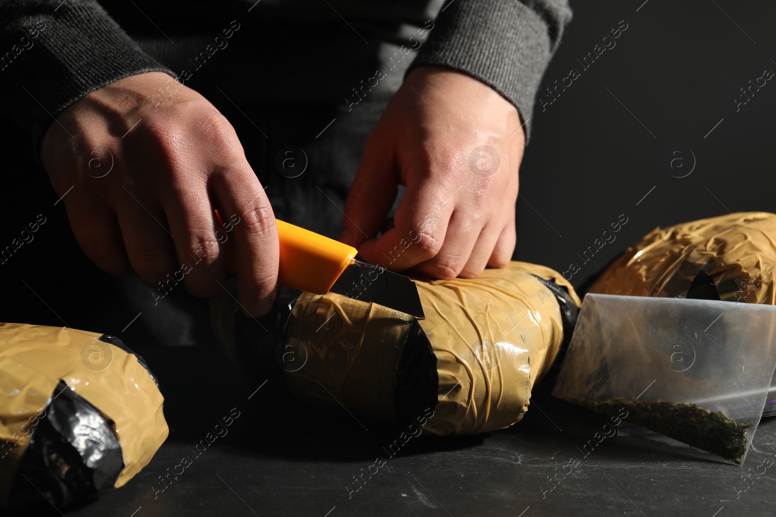 Photo of Smuggling and drug trafficking. Man opening package of narcotics with box cutter at dark table, closeup