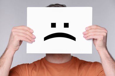 Man hiding behind sheet of paper with sad face on grey background