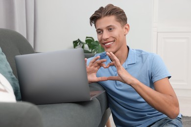 Photo of Happy young man having video chat via laptop and making heart with hands on sofa indoors. Long-distance relationship