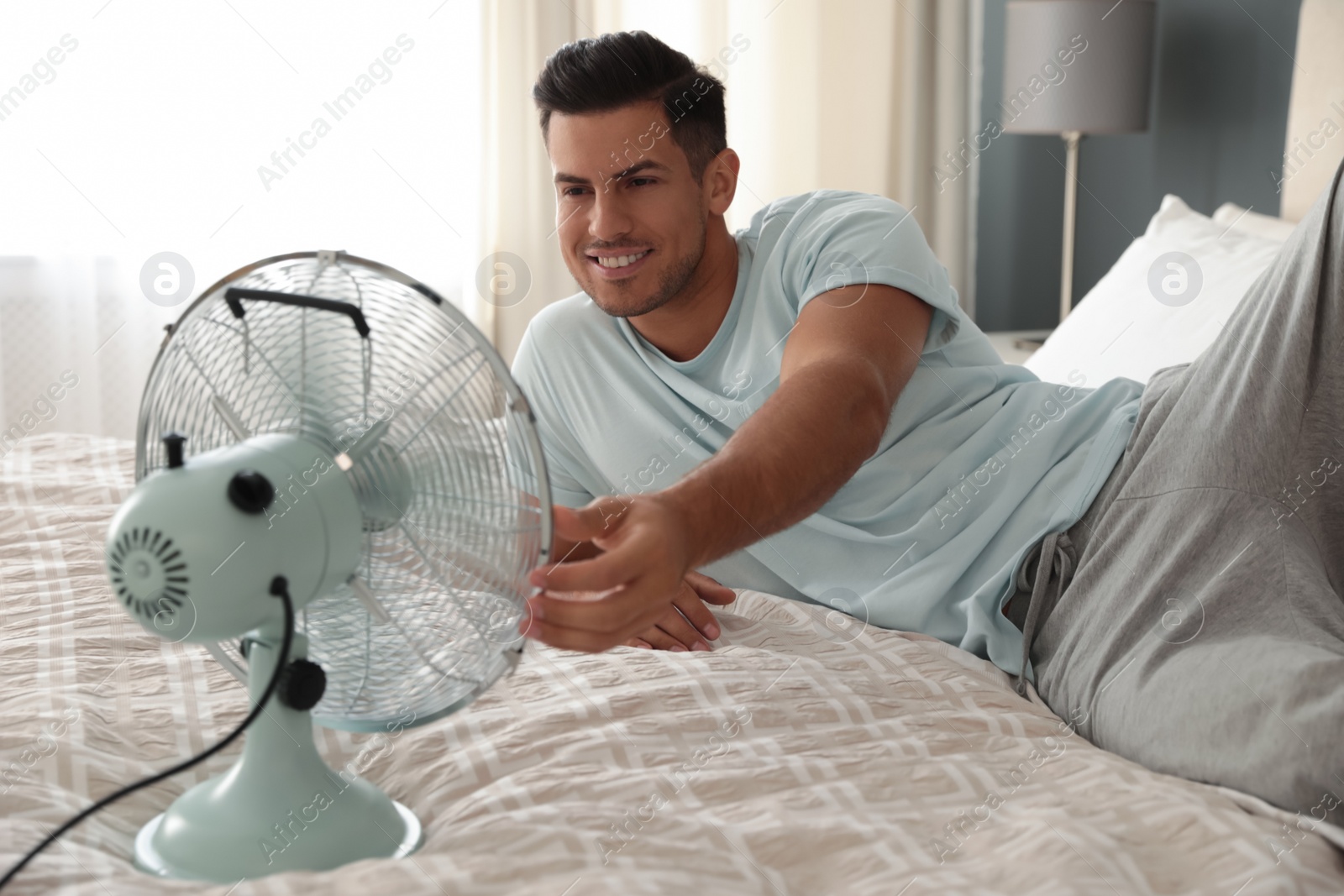 Photo of Man enjoying air flow from fan on bed in room. Summer heat