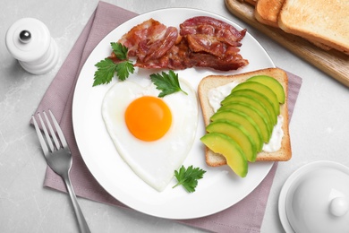 Photo of Romantic breakfast with fried bacon, heart shaped egg and avocado toast on light grey table, flat lay. Valentine's day celebration