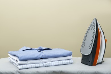 Photo of Modern iron and stack of clean clothes on board against beige background. Space for text