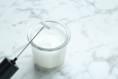 Mini mixer (milk frother) and whipped milk in glass on white marble table, closeup. Space for text